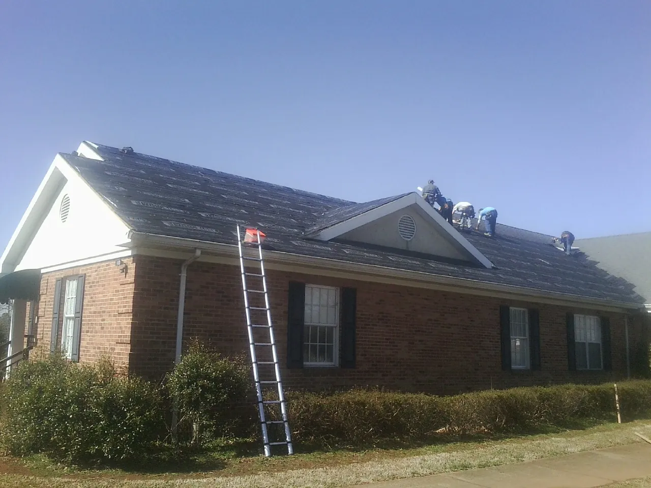 Crew tearing shingles off a brick ranch roof with a ladder set — Emergency Roof Repair work by our crew in Flom