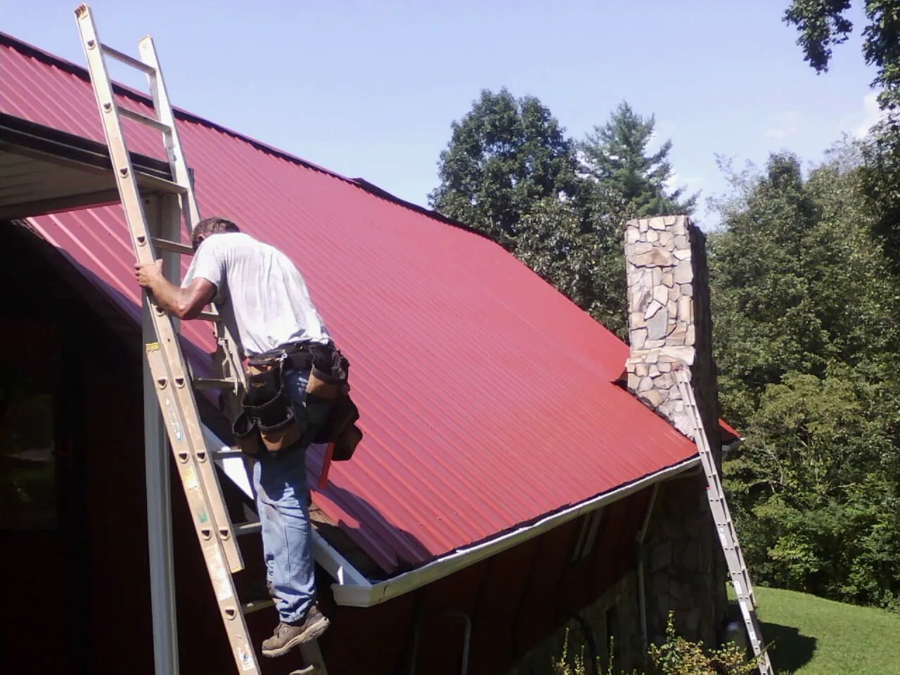 Roofer on a ladder beside a red metal roof and stone chimney — Soffit & Fascia Repair by Roofing Brothers in Flom